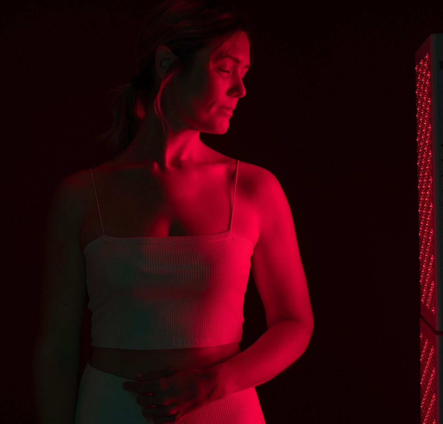 A woman in a white sleeveless top stands in a dark room, illuminated by red light from a vertical LED panel on her right. She looks towards the light, with her face and body bathed in a warm red glow.