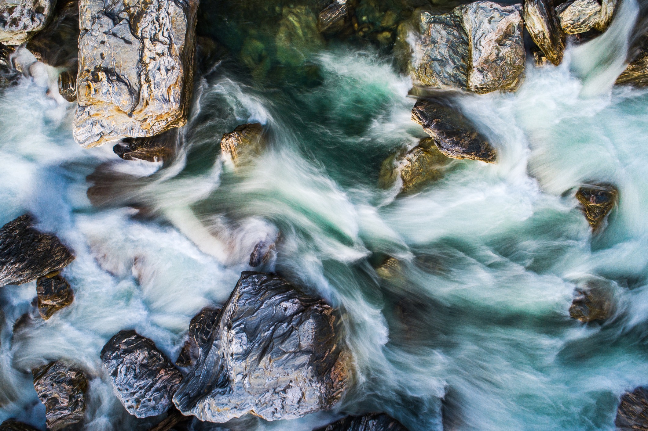 Aerial view of fast-moving, white and turquoise river water flowing around large, rugged rocks, creating swirling patterns and a sense of motion.
