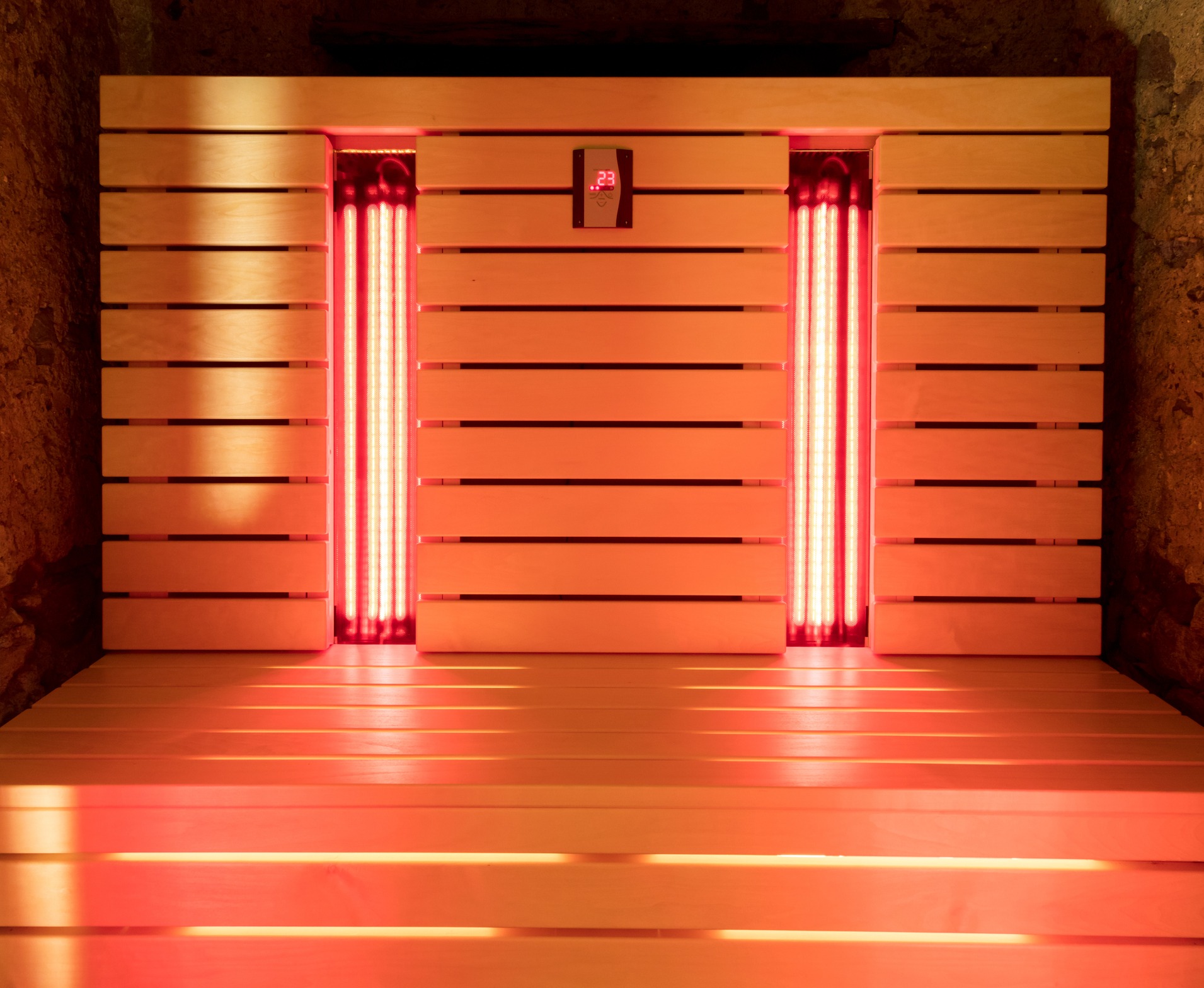 A modern wooden infrared sauna with horizontal slats, glowing red heat lamps, and a digital temperature display on the wall, emitting a warm amber light.