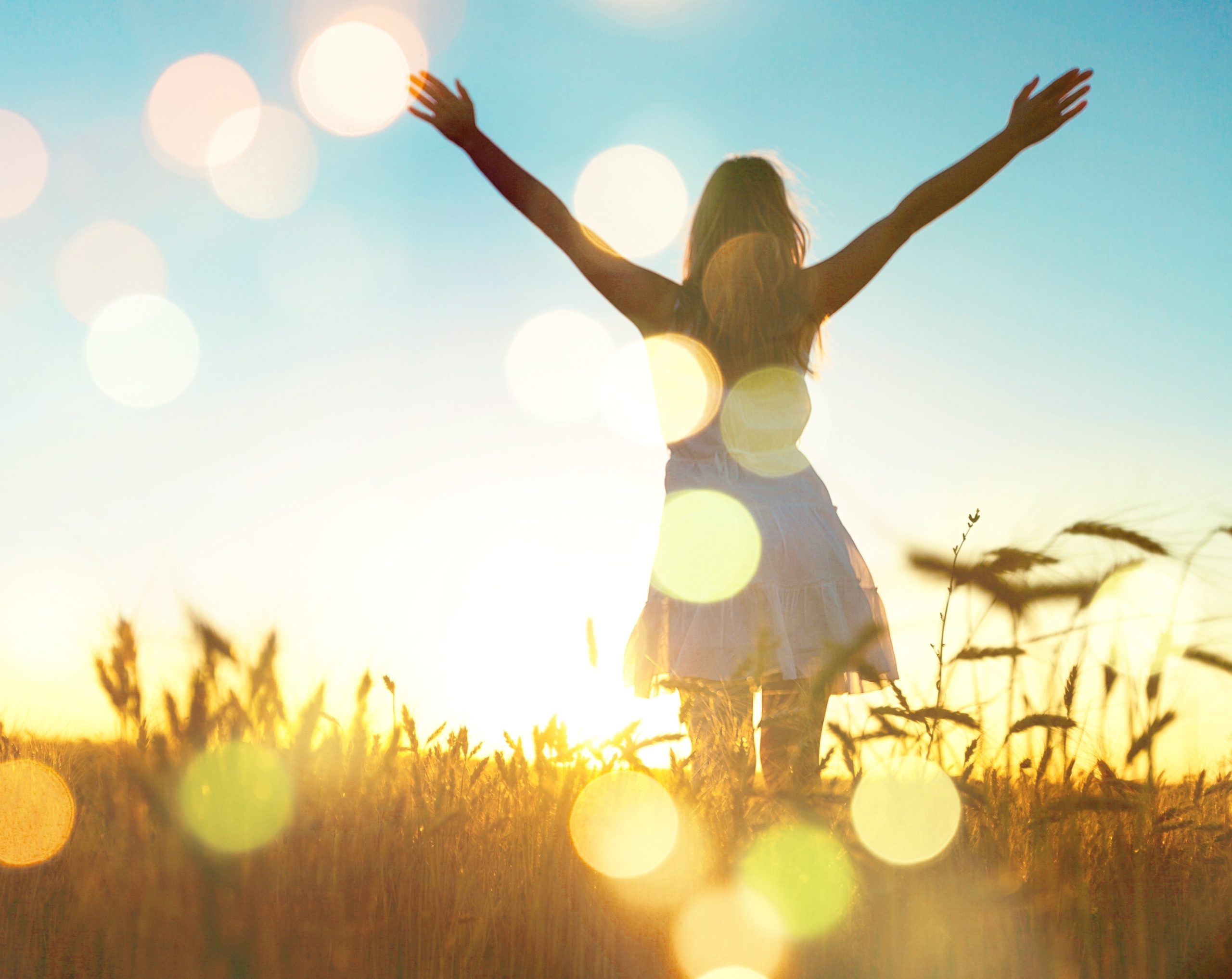 A woman stands in a sunlit field with her arms raised, facing the sunrise. She wears a white dress, and golden light and lens flares create a warm, uplifting atmosphere.