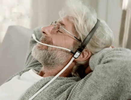 An older man with gray hair and a beard is relaxing with his hands behind his head, eyes closed, and wearing a headset with a microphone.