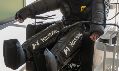 COMPRESSION SLEEVES A person sits in a chair wearing large black Normatec compression sleeves on their arms and legs, likely undergoing a recovery or therapy session. Only the upper torso, arms, and legs are visible.