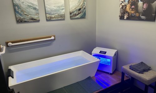 CRYOTHERAPY A clean, modern ice bath room with a large white tub filled with water, an ice machine nearby, a towel on a bench, and wall art including three wave paintings and a photo of three dogs.