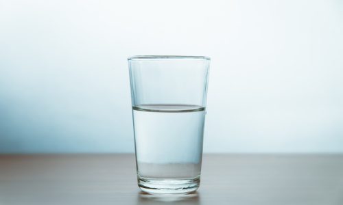 Glass Of Water On Table Against Wall A clear glass filled halfway with water sits on a smooth, light-colored surface with a plain, softly lit background.