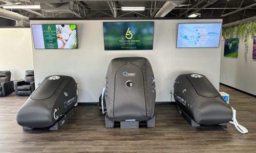 HYPERBARIC CHAMBERS Three black hyperbaric oxygen therapy chambers are positioned in a row against a white wall, with three digital screens mounted above them displaying information and graphics in a modern wellness center setting.