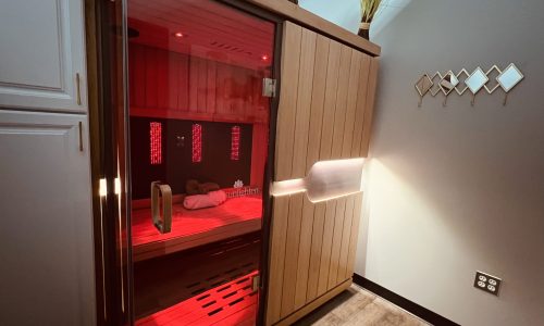 INFRARED SAUNA A modern wooden infrared sauna with a glass door emits a red glow. Towels are inside, and decorative grass and geometric wall art are visible nearby in a spa-like room.