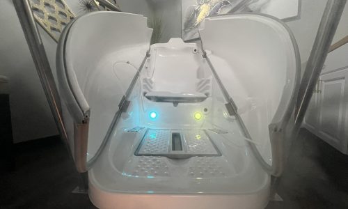 OZONE Wide-angle view of an open futuristic spa pod with glowing green and blue lights inside, surrounded by mist, in a modern, softly lit room with contemporary decor.