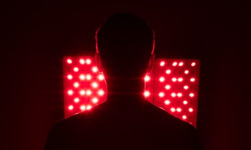 RED LIGHT THERAPY A person’s silhouette faces two panels of bright red lights in a dark room, creating a dramatic, high-contrast scene.