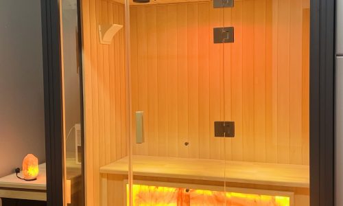 SALT THERAPY A modern glass-enclosed sauna with warm interior lighting, wooden bench, and Himalayan salt panel. Potted plants sit on top of the sauna and a small salt lamp glows on a table nearby.
