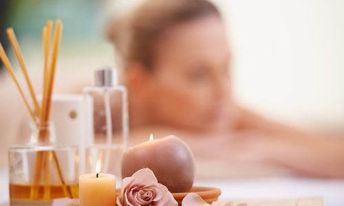 Basking,In,The,Afterglow,Of,Her,Massage.,Ropped,Shot,Of A relaxing spa setting with scented candles, a pink rose, rose petals, and a reed diffuser in the foreground. In the blurred background, a person lies face down, suggesting a calming environment.
